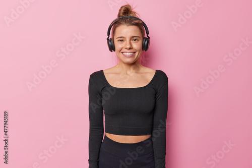 Waist up shot of active happy young sportswoman in black tracksuit listens favorite song via wireless headphones exercises regularly indoor isolated over pink background. Workout with music.