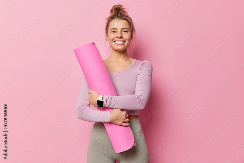 Fototapeta premium Pleased fitness trainers going to conduct yoga class embraces rolled karemat smiles gladfully has combed hair dressed in activewear uses smartwatch isolated over pink backgound. Time for training