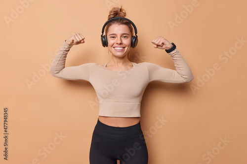 Horizontal shot of active slim young woman raises arms shows biceps after workout dressed in sportswear has happy expression listens music in wireless headphones isolated over beige background