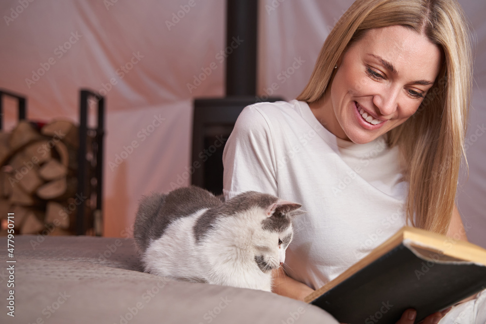 Pleased woman reading book with cat by her side Pleased woman reading book with cat by her side