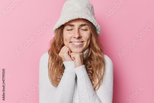Portrait of happy woman closes eyes from satisfaction keeps hands under chin smiles jofuly has dimples on cheeks wears winter hat and white sweater isolated over pink background. Pleasant feelings photo