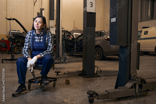 Joyous lady in overalls resting on chair at repair shop
