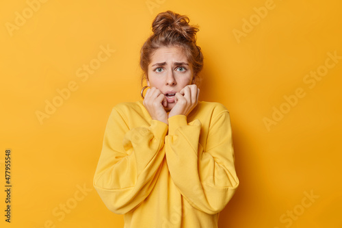 Concerned anxious European woman trembles from fright grabs face stands shocked indoor notices something horrified dressed in jumper isolated over yellow background. Human reactions concept. photo