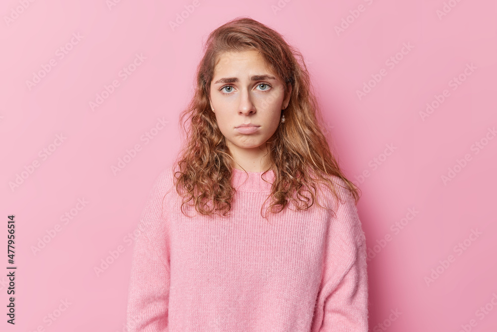 Upset gloomy blue eyed woman has pity look feels discontent and offended looks dissatisfied pouts lips dressed in casual jumper isolated over pink background expresses sadness. Negative emotions