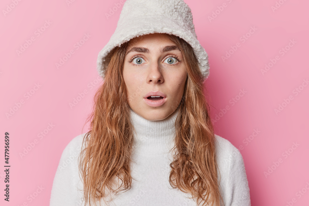 Indoor shot of impressed wondered woman holds breath stares with widely opened eyes holds breath hearss shocking newss wears winter hat and sweater isolated over pink background. Omg concept Indoor shot of impressed wondered woman holds breath stares with widely opened eyes holds breath hearss shocking newss wears winter hat and sweater isolated over pink background. Omg concept