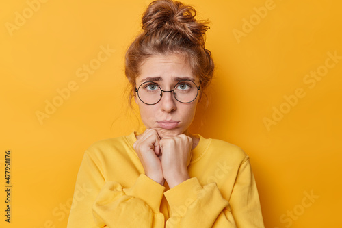 Portrait of sad offended woman keeps hands under chin feels displeased has gloomy sulking expression wears round spectacles jumper isolated over vivid yellow background thinks of something upsetting
