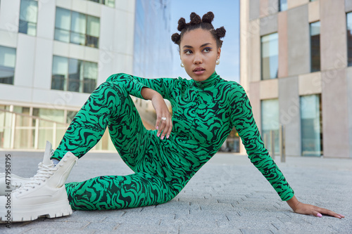 Thoughtful pretty woman with trendy hairstyle wears green bodysuit and white boots poses in urban setting during daytime being deep in thoughts admires something. People and lifestyle concept photo