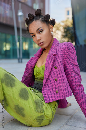 Vertical shot of beautiful dark skinned serious woman with hair buns dressed in fashionable clothes poses outdoors against urban background. Street photography. People style and lifestyle concept
