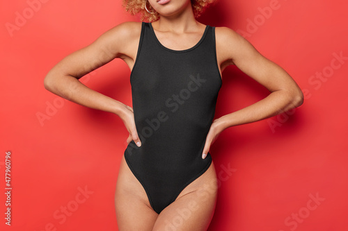 Cropped shot of slim woman in black bodysuit keeps hands on waist has fit body slender legs perfect figure isolated over vivid red background. Unrecognizable female model in good physical shape photo