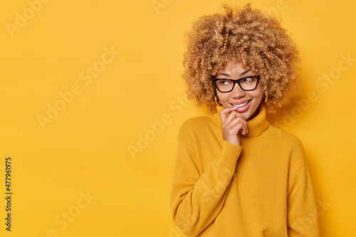 Horizontal shot of dreamy lovely young woman keeps finger on lips looks away with curious happy expression dressed in comfortable jumper and spectacles isolated over yellow background copy space © Wayhome Studio