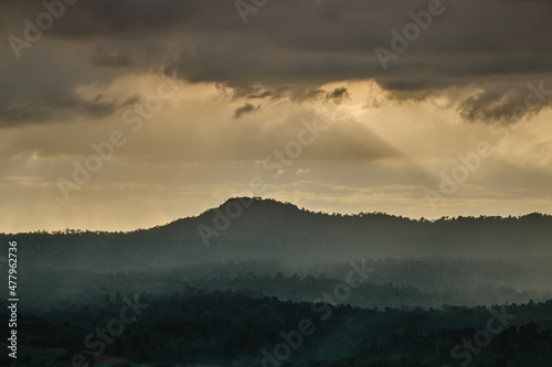 Sunset over the mountains in cloudy sky with sun rays down to the hills with green forest and agricultural field