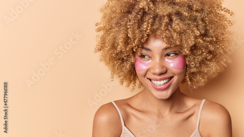 Positive woman with curly bushy hair looks away has happy expression smiles toothily has healthy clean skin well cared complexion applies pink hydrogel patches under eyes poses indoor blank space