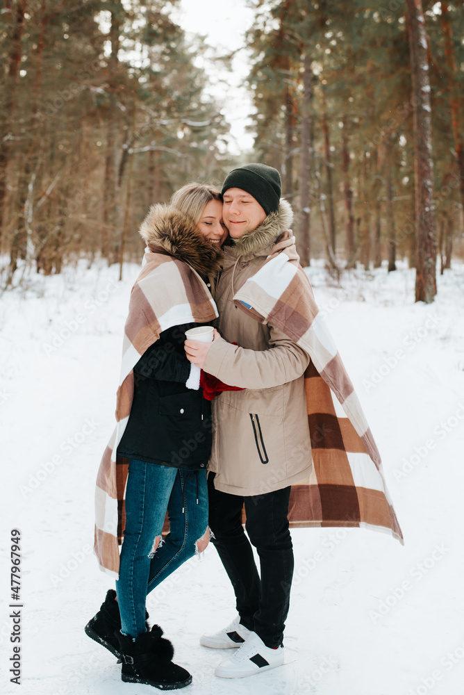 Happy young couple of lovers covered with blanket hug during joint romantic walk in winter snowy forest, outdoors. Caucasian hipster family enjoying outdoor vacation together