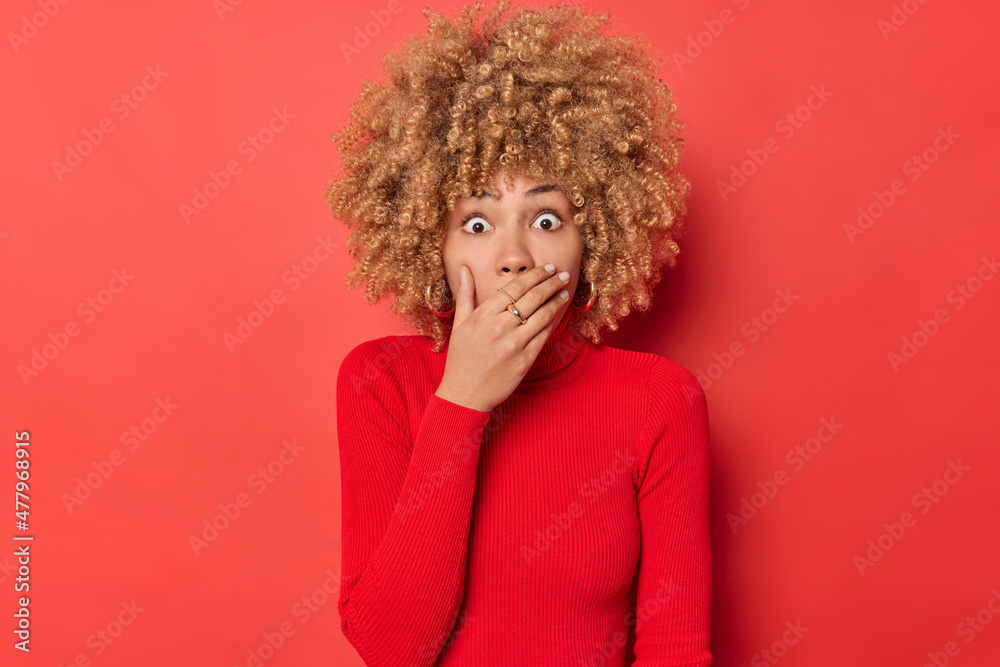 Shocked curly haired woman gasps from wonder covers mouth with hand stares bugged eyes witnesses terrible scene wears casual turtleneck isolated over red background. Human reactions concept. Shocked curly haired woman gasps from wonder covers mouth with hand stares bugged eyes witnesses terrible scene wears casual turtleneck isolated over red background. Human reactions concept.