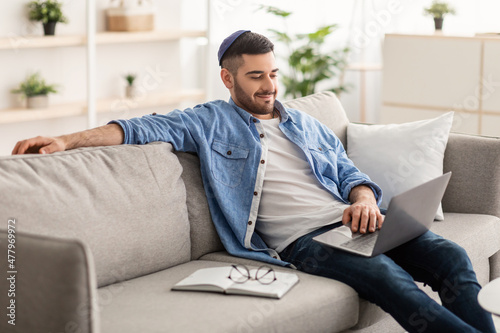Cheerful male jew working on laptop at home on sofa