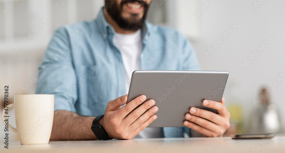 Work remotely from home. Unrecognizable young bearded man holding digital tablet, sitting at table in kitchen, crop Work remotely from home. Unrecognizable young bearded man holding digital tablet, sitting at table in kitchen, crop