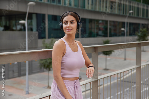 Outdoor shot of thoughtful healthy sportswoman listens music in headphones wears top and shorts checks time on smartwatch poses at bridge against city background concentrates away into distance Free Download Outdoor shot of thoughtful healthy sportswoman listens music in headphones wears top and shorts checks time on smartwatch poses at bridge against city background concentrates away into distance - GFXLoad
