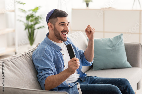 Happy jewish guy spending weekend watching tv shaking clenched fists