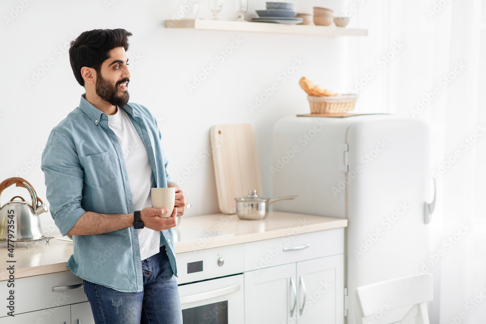 Start of the day. Happy arab man drinking morning coffee in kitchen, standing with cup of hot drink, empty space Start of the day. Happy arab man drinking morning coffee in kitchen, standing with cup of hot drink, empty space
