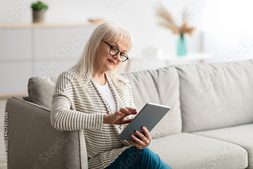 Mature lady in glasses using digital tablet sitting on couch