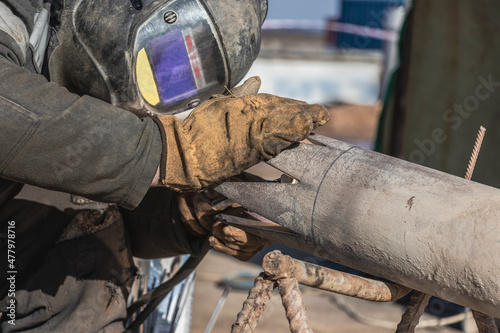 A welder in work clothes is welding a pipe at a construction site. Close-up.