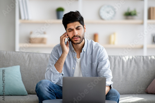 Remote business. Concerned arab freelancer talking on cellphone and using laptop computer, sitting on sofa at home © Prostock-studio