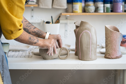 Ceramics workshop. Cropped photo of woman artisan making earthenware mug, working with clay during pottery masterclass, tattooed hands of female ceramist sculpting or modeling handicrafter kitchenware photo