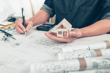 Architect engineer hands hold and show mini house model and use pen drawing on bueprint. House planning design and construction concept.