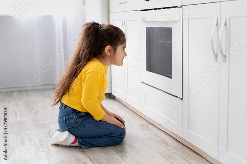 Cute little girl sitting near oven in kitchen and looking inside