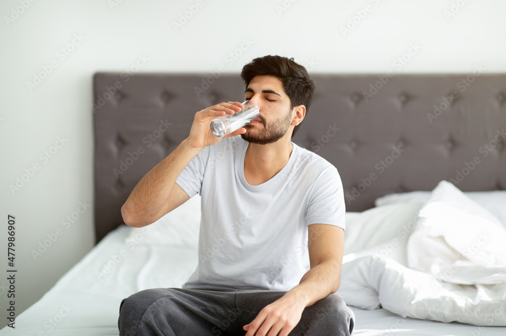 Starting new day with fresh aqua. Arab man taking glass of water sitting on bed in the morning Starting new day with fresh aqua. Arab man taking glass of water sitting on bed in the morning