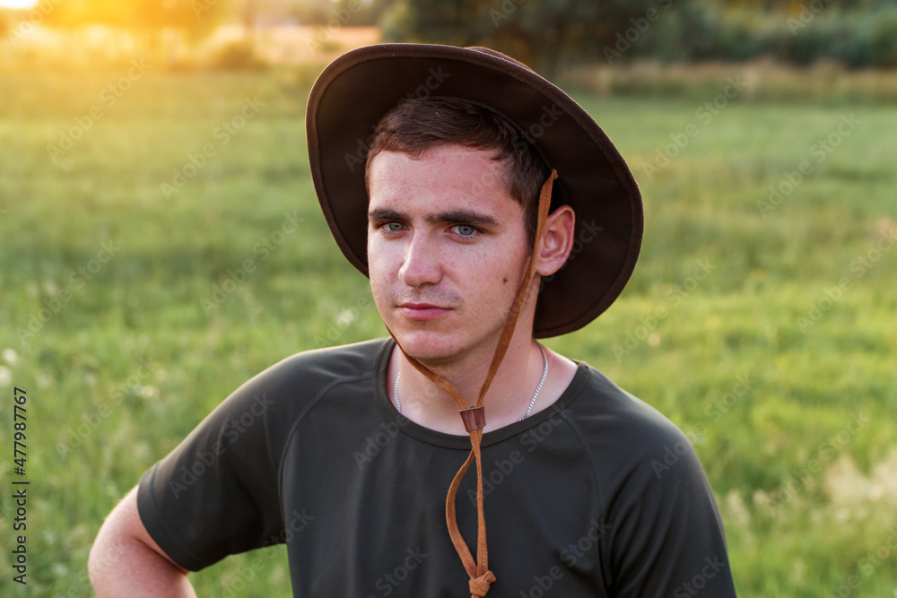 Obraz premium Young man farmer in cowboy hat at agricultural field on sunset with sun flare. Closeup portrait of millennial man with hat, standing on nature background, outdoors. Countryman, rancher, cowboy