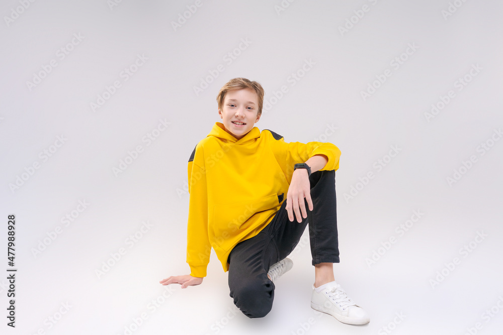 Young handsome happy smiling guy teenager in black jeans, light yellow sweatshirt with a hood, rejoices while sitting posing on a light background. The concept of emotions, good mood Young handsome happy smiling guy teenager in black jeans, light yellow sweatshirt with a hood, rejoices while sitting posing on a light background. The concept of emotions, good mood