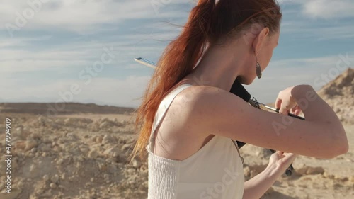 Young pretty slender red-haired woman with long hair in a white dress is playing the violin on a hill on a sunny day. In the background we can see the sea and the city