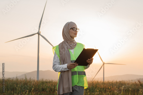 Charming muslim woman in hijab and eyeglases holding clipboard in hands while posing among field with wind turbines. Portrait of female inspector on eco farm during summer sunset. photo