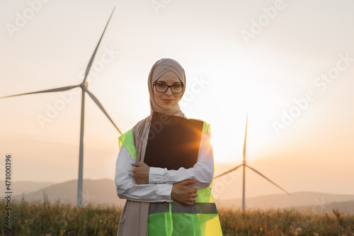 Charming muslim woman in hijab and eyeglases holding clipboard in hands while posing among field with wind turbines. Portrait of female inspector on eco farm during summer sunset. photo