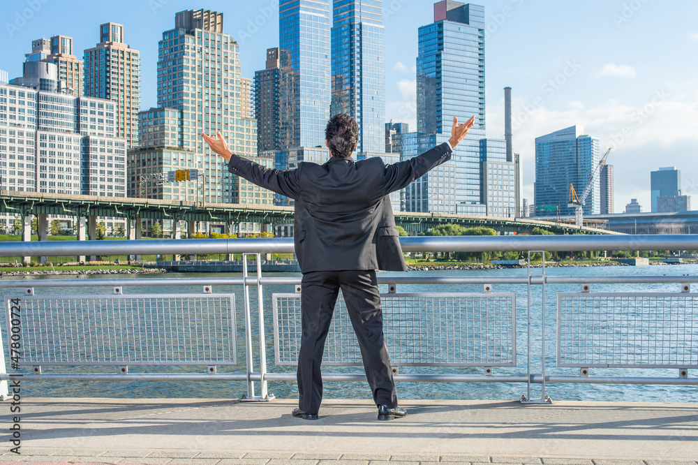 Facing dramatic high buildings of New York City, a middle age businessman is raising both arms, a symbol of success.. Facing dramatic high buildings of New York City, a middle age businessman is raising both arms, a symbol of success..