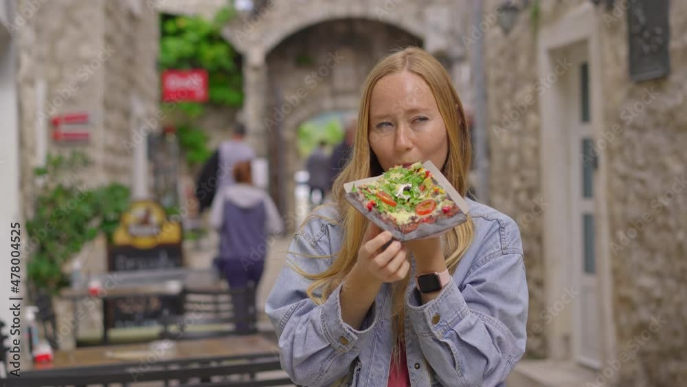 A young woman eats a delicious pizza on black bread standing on a street of an old European town A young woman eats a delicious pizza on black bread standing on a street of an old European town