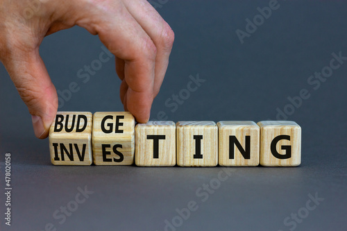 Budgeting or investing symbol. Businessman turns cubes and changes the word investing to budgeting. Beautiful grey table, grey background, copy space. Business and budgeting or investing concept.