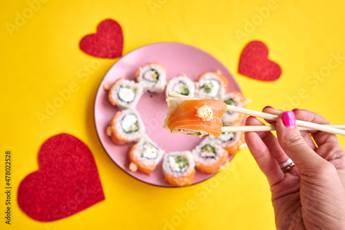 Sushi rolls with salmon in a heart shape and a woman's hand with chopsticks on a yellow background.Creative sushi concept for Valentines day or romantic dinner 