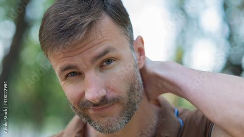 In the park, a handsome man with a beard looks skeptically at the camera.