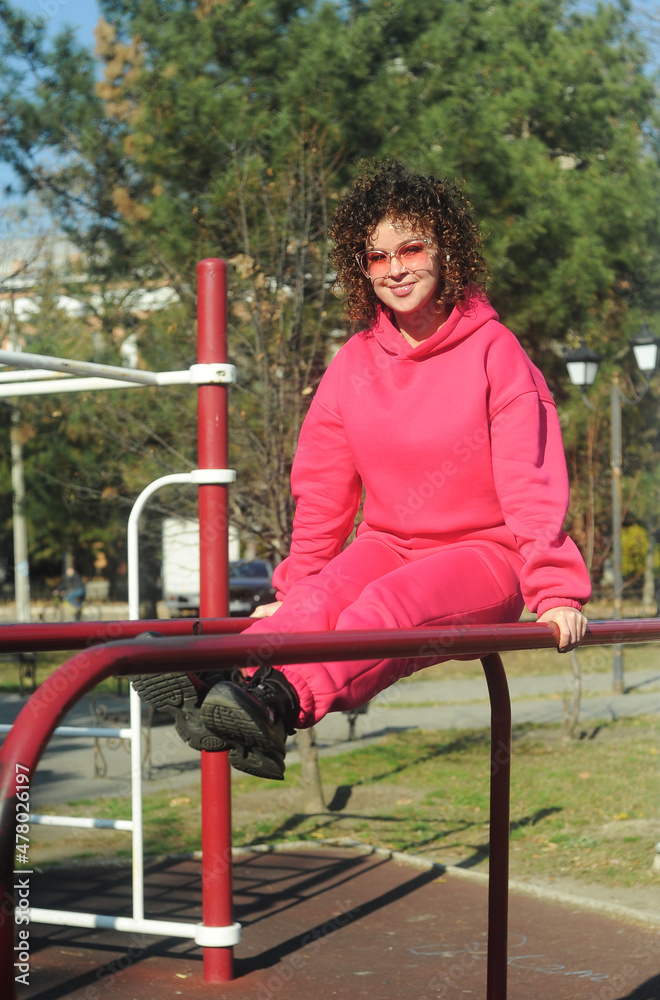 Cute curly haired woman in pink sportswear makes exercises on playground