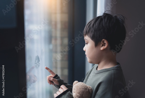 Portrait of school boy standing next to window and using his finger drawing or writing on window glass, Side view of child hand drawing on glass, Toddler boy looking at him self through window.