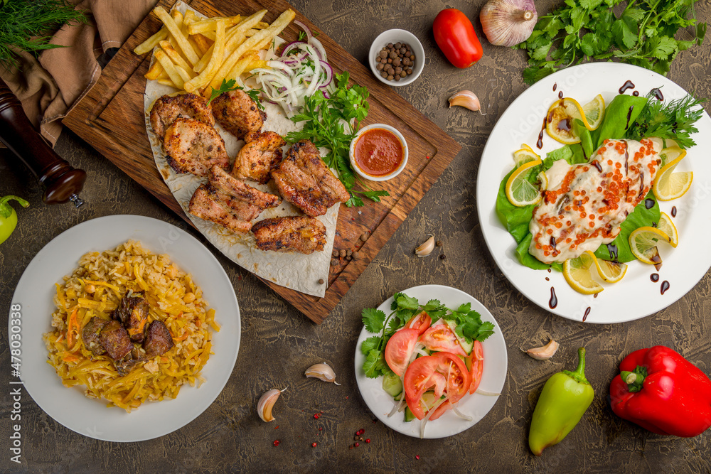 Kebab of pork, steak of salmon on creamy sauce with red caviar, vegetable salad and pilaf with beef top view on brown table Kebab of pork, steak of salmon on creamy sauce with red caviar, vegetable salad and pilaf with beef top view on brown table