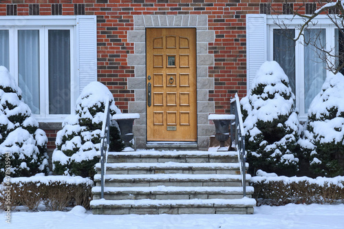 Traditional brick house with wood grain front door and snow covered shrubbery Fototapeta