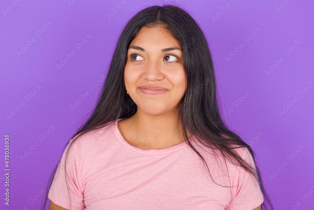 Hispanic brunette girl wearing pink t-shirt over purple background looking aside into empty space thoughtful Hispanic brunette girl wearing pink t-shirt over purple background looking aside into empty space thoughtful