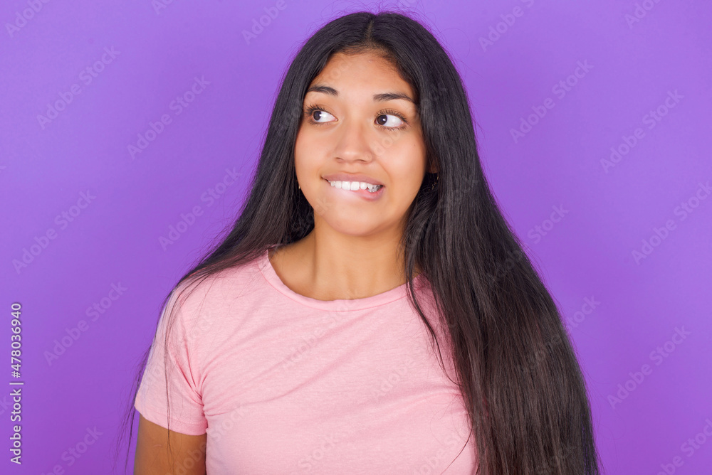 Fototapeta premium Amazed Hispanic brunette girl wearing pink t-shirt over purple background bitting lip and looking tricky to empty space.