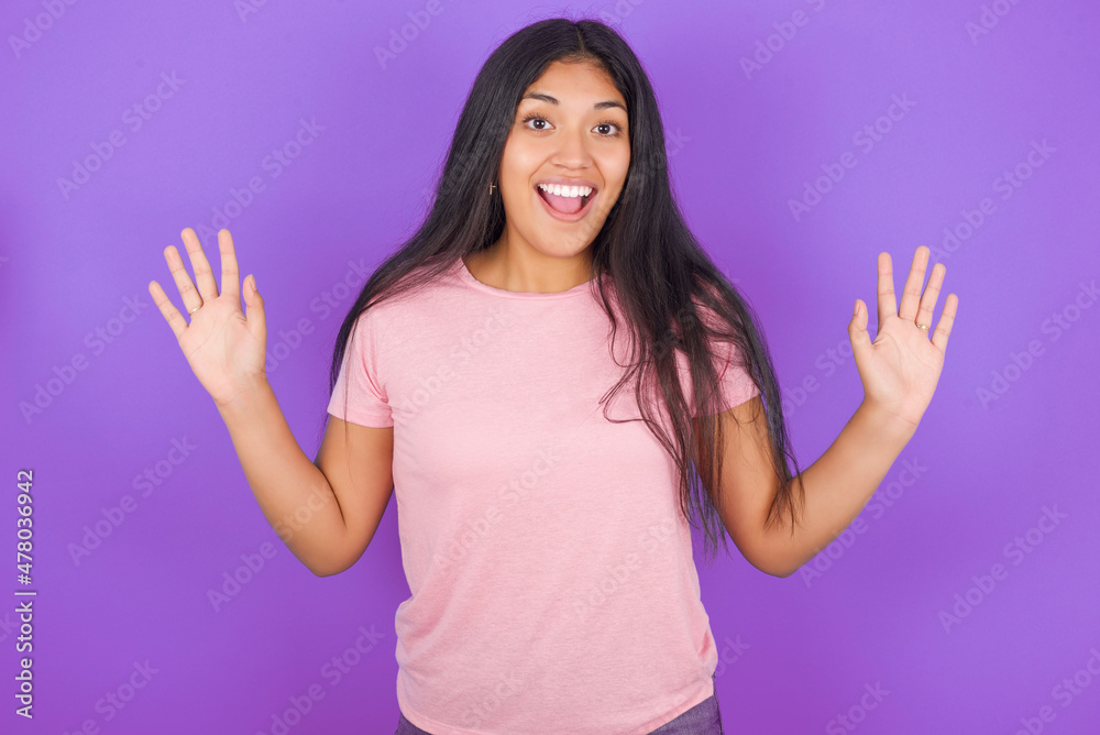Optimistic Hispanic brunette girl wearing pink t-shirt over purple background raises palms from joy, happy to receive awesome present from someone, shouts loudly, Excited model screaming. Optimistic Hispanic brunette girl wearing pink t-shirt over purple background raises palms from joy, happy to receive awesome present from someone, shouts loudly, Excited model screaming.