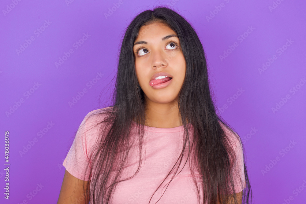 Hispanic brunette girl wearing pink t-shirt over purple background showing grimace face crossing eyes and showing tongue. Being funny and crazy Hispanic brunette girl wearing pink t-shirt over purple background showing grimace face crossing eyes and showing tongue. Being funny and crazy