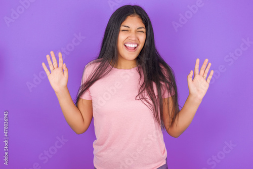 Emotive Hispanic brunette girl wearing pink t-shirt over purple background laughs loudly, hears funny joke or story, raises palms with satisfaction, being overjoyed amused by friend