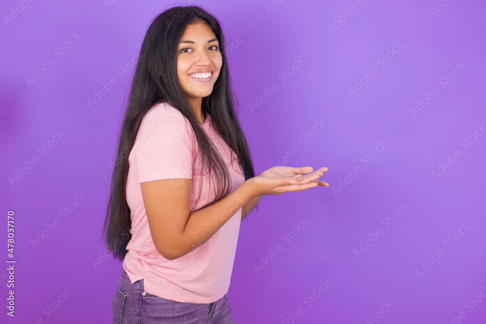 Hispanic brunette girl wearing pink t-shirt over purple background pointing aside with hands open palms showing copy space, presenting advertisement smiling excited happy Hispanic brunette girl wearing pink t-shirt over purple background pointing aside with hands open palms showing copy space, presenting advertisement smiling excited happy